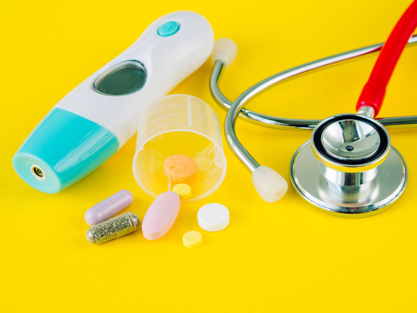 Stethoscope and pills with an electronic thermometer on yellow background - Photo, Image