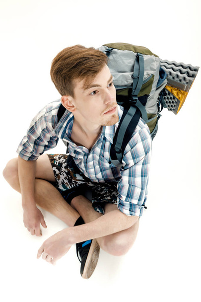 Funny Young tourist is sitting with backpack on white background. traveler is preparing for hike - Foto, imagen
