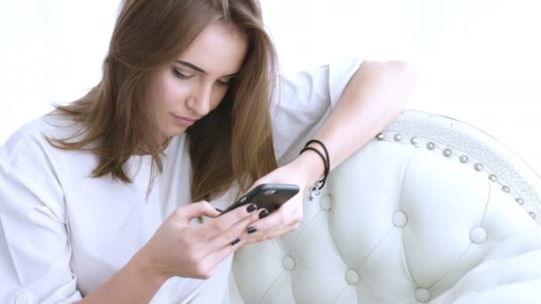 Young beautiful girl texting on mobile phone sitting on the sofa. Woman using app on smartphone - Materiaali, video