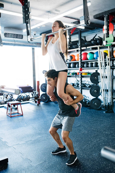 Woman in gym doing pull ups, man giving her piggyback. - Zdjęcie, obraz