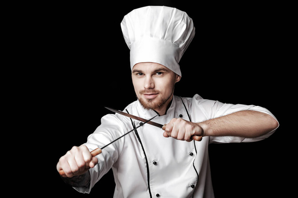 Young bearded chef in white uniform holding two knives on black background - Valokuva, kuva