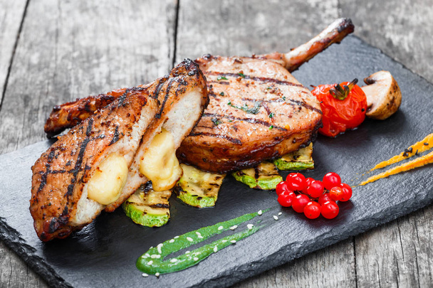 Roasted Pork steak on bone stuffed with cheese, grilled vegetables and berries on stone slate background on wooden background close up. Hot Meat Dishes. Top view with copy space - Photo, Image