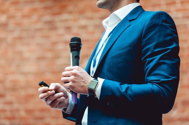 Business and speech topic: Man in a blue suit holding a gray microphone a on a orange bricks background - Photo, Image