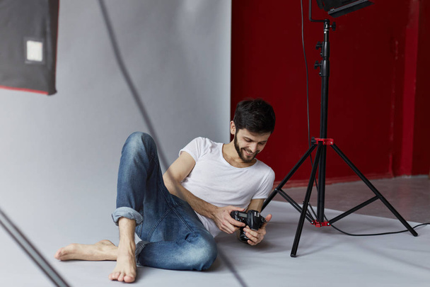 Studio portret przystojny kaukaski mężczyzna pozowanie na szarym tle. Młody atrakcyjny Brodaty fotograf z aparatu w rękach jest oglądanie zdjęć.  - Zdjęcie, obraz
