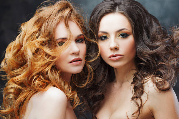 Two charming beauties with luxurious curly hair. Brunette and redhead. Studio portrait on the dark background. - Zdjęcie, obraz