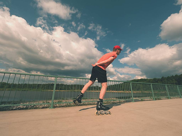 Vista traseira para patinador em linha em camiseta vermelha e calças pretas patinando na ponte. Patinação ao ar livre em linha
  - Foto, Imagem