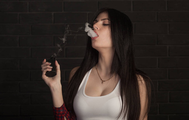 Jeune belle femme brune fumant, vaping e-cigarette avec de la fumée sur fond noir
. - Photo, image
