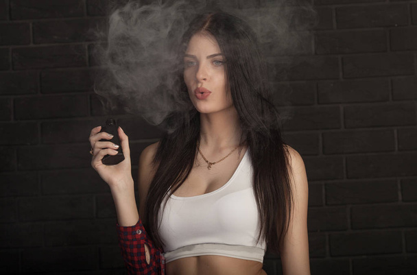 Young beautiful brunette woman smoking, vaping e-cigarette with smoke on black background. - Foto, afbeelding