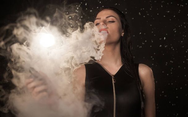 Young beautiful woman vaping e-cigarette. Water flowing on woman face. - Photo, Image
