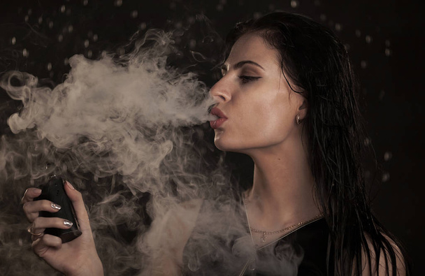 Young beautiful woman vaping e-cigarette. Water flowing on woman face. - Photo, image