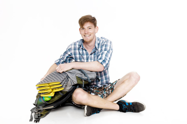 Young smiling tourist is sitting with backpack on white background. traveler is preparing for hike - Foto, afbeelding