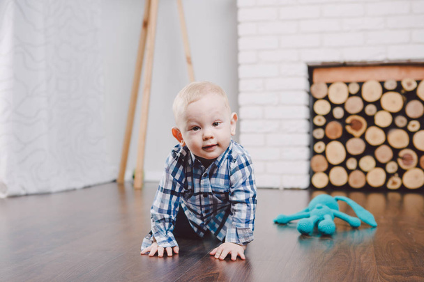 A small child, a boy sits on a wooden floor in a bright room for a year. Dressed in blue jeans and a shirt. Family concept - Photo, Image