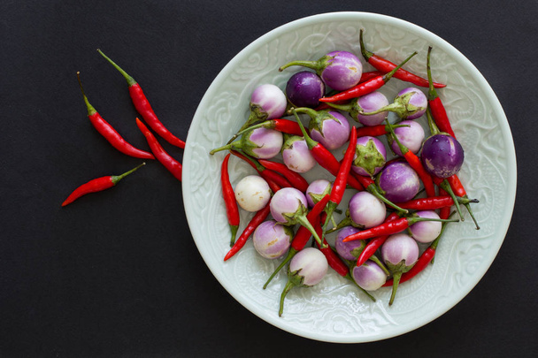 lila thailändische Auberginen und scharfe Chilischoten auf einem grünen Teller und daneben Salzkristalle auf schwarzem Hintergrund. Asiatisches Essen. - Foto, Bild