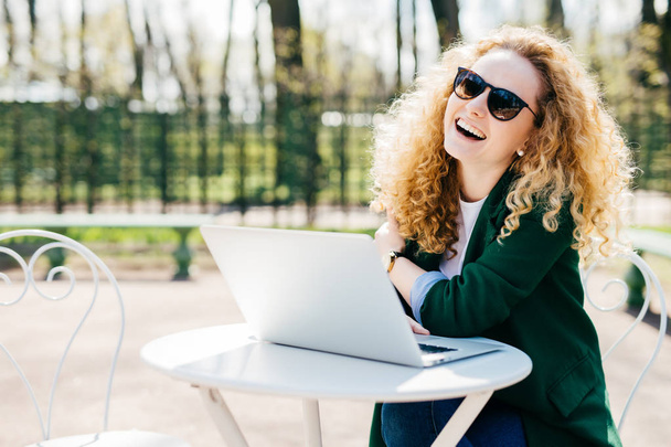 Boldog szép szőke nő visel, napszemüveget és zöld kabát rendelkező többi ülő-on fehér iskolapad használ a laptop kommunikál barátaival online park, göndör haj. Emberek, érzelmek fogalma - Fotó, kép