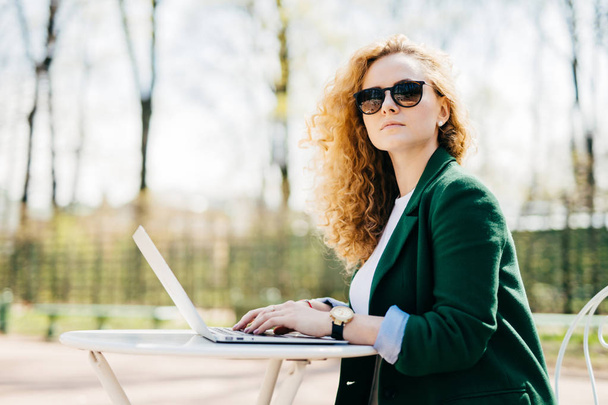 Personas, ocio, tecnología y comunicación. Hermosa mujer de negocios con gafas de sol y chaqueta elegante usando computadora portátil para hacer conversación con sus socios sentados de lado en el escritorio blanco
 - Foto, imagen