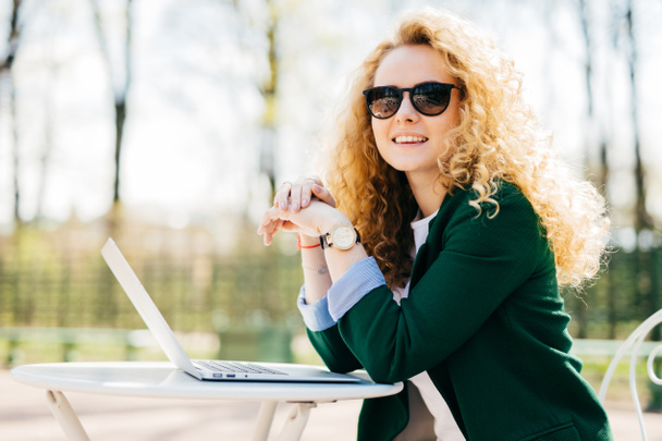 Beautiful young student female with beautiful curly blonde hair elegant dressed using generic laptop computer for writing essay having rest outdoors having charming smile looking into distance - Foto, immagini
