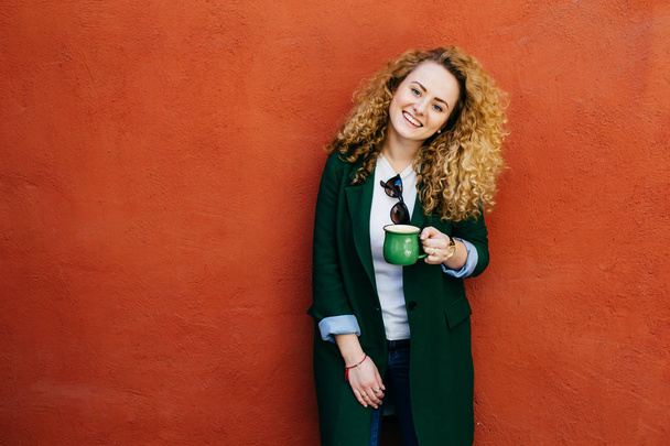 Довольно кудрявая женщина в стильной одежде, держа чашку кофе с широкой улыбкой на лице, глядя прямо в камеру изолированы над оранжевой стеной с копией пространства для текста или рекламного содержания
 - Фото, изображение