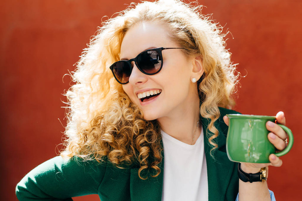 Lateralmente primo piano di donna attraente che indossa tonalità eleganti e giacca in possesso di una tazza di tè verde con sorriso sincero sul suo viso guardando da parte godendo la sua vita isolata su sfondo arancione
 - Foto, immagini