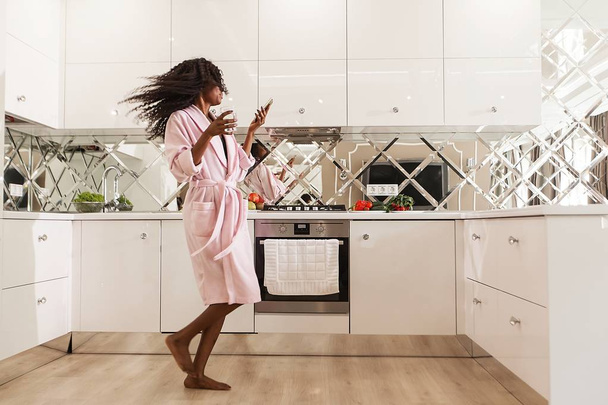 Slender black woman dancing in the kitchen with the telephone and glass of water in her hands. - Foto, imagen