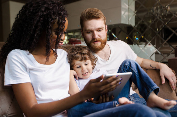 Happy family photo. Black woman with white man and child sitting on the sofa and looking at the tablet screen. - Foto, afbeelding