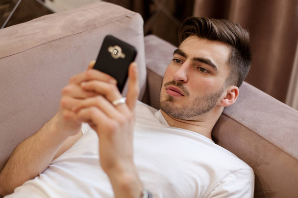 Handsome young man lying on the sofa and looking at the phone screen. - Фото, изображение