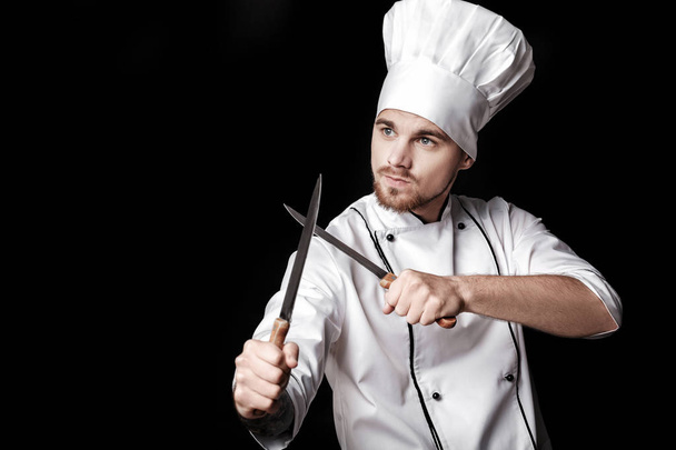 Young bearded man chef In white uniform holds Two knives on  black background - Fotó, kép