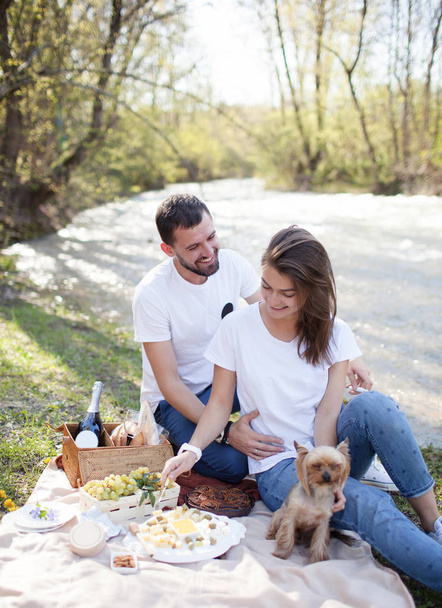 Picnic on the grass near river on lazy summer day. Young smiling couple with small dog relaxing on nature - Photo, image