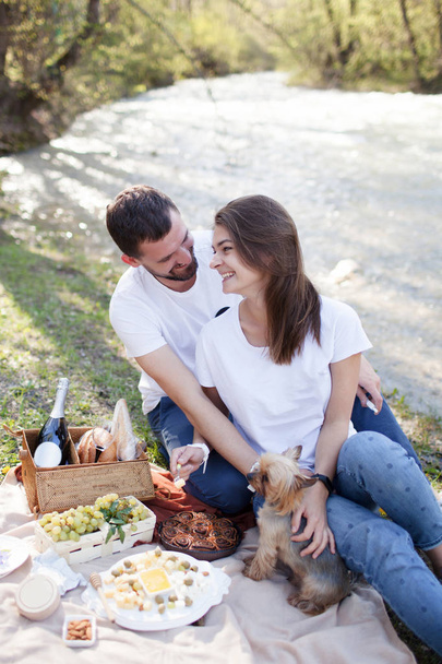 Picnic on the grass near river on lazy summer day. Young smiling couple with small dog relaxing on nature - Foto, Imagen