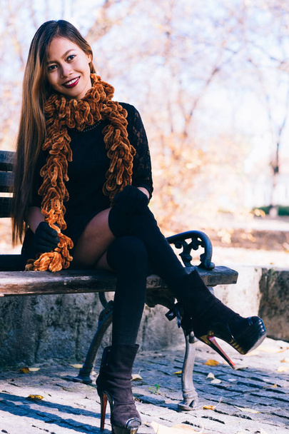 Young beautiful girl in black dress with brown scarf sitting on the bench - 写真・画像