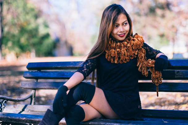 Young beautiful girl in black dress with brown scarf sitting on the bench - 写真・画像