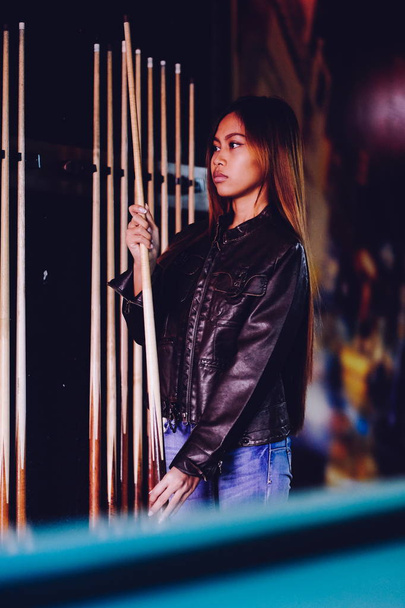 Young beautiful girl wearing leather jacket in a billiard club, with cue stick preparing for the game - Φωτογραφία, εικόνα