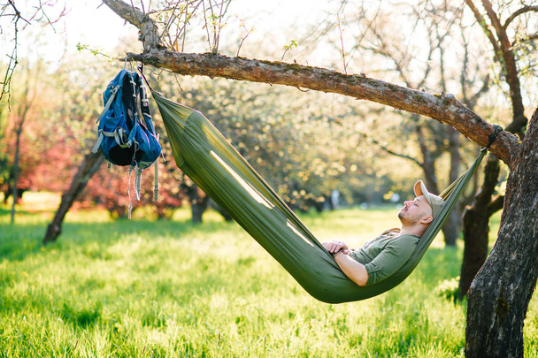 Happy traveler bearded hipster man in green hat lying in hummock on apple tree in blooming garden in summer sunny day on vacation. Weekend and holiday. Tourism. Leisure. Dreaming. Fragrance in air. - Photo, image