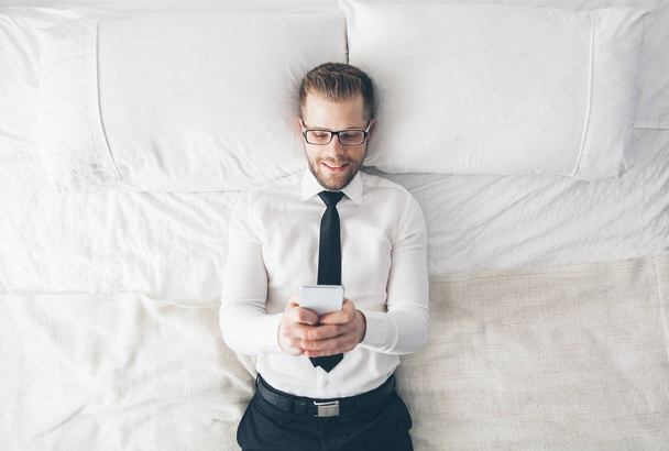 Pohled shora. Pohledný podnikatel s brýlemi ležící na posteli SMS ze svého mobilního telefonu - Fotografie, Obrázek