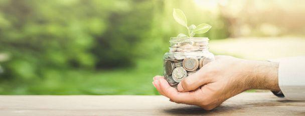 Plant growing from money (coins) in the glass jar held by a man's hands  - business and financial metaphor concept, web banner with copy space - Photo, Image