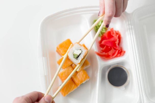 Hand houden met stokjes, broodjes, sushi roll. Chopsticks, gember, sojasaus in container levering. - Foto, afbeelding