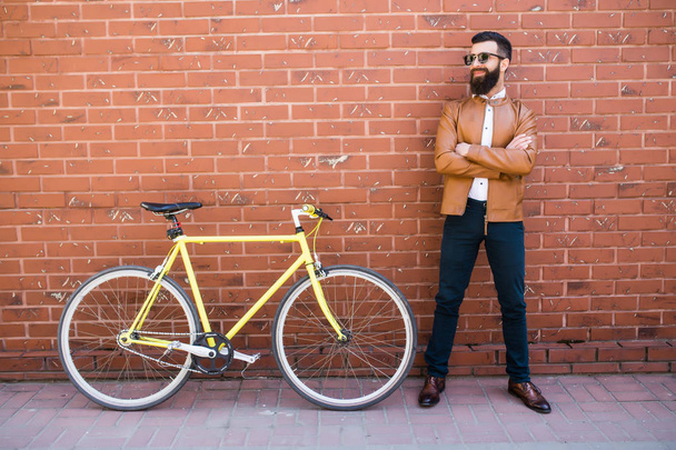 Handsome young bearded man while standing near his bicycle against the brick wall - Фото, изображение