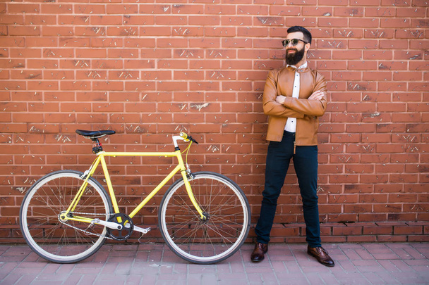 Handsome young bearded man while standing near his bicycle against the brick wall - Foto, imagen
