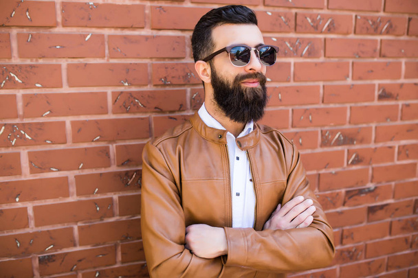 Portrait of a brutal bearded man on brick wall background. Young stylish hipster posing in the street. - 写真・画像