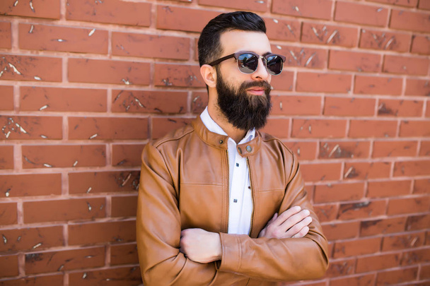 tuğla duvar arka plan üzerinde acımasız bir sakallı adam portresi. Genç şık hipster sokağın ortasında poz. - Fotoğraf, Görsel