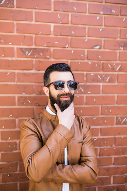 портрет жестокого бородатого человека на фоне кирпичной стены. Молодой стильный хипстер позирует на улице
. - Фото, изображение