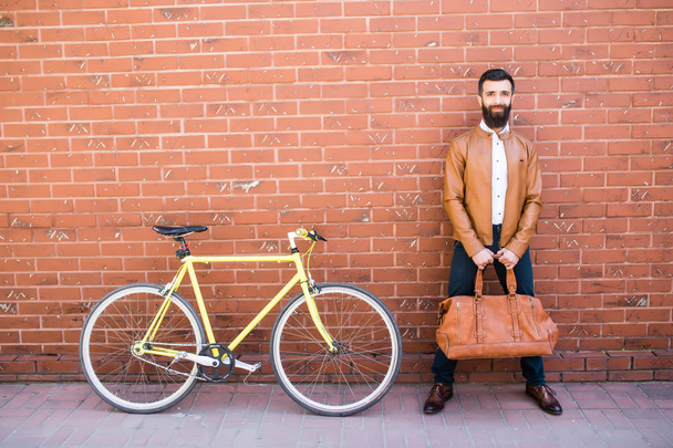 Nuori tyylikäs mies parta tiili tausta seisoo lähellä pyörä laukku käsissä
 - Valokuva, kuva