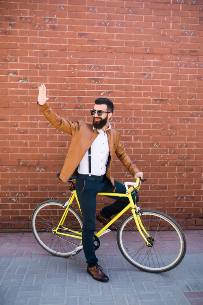 счастливый молодой хипстер с фиксированной передачей велосипед размахивая рукой на городской улице
 - Фото, изображение