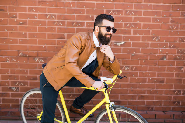 Homem bonito jovem com uma barba contra a parede de tijolo senta-se em uma bicicleta
 - Foto, Imagem