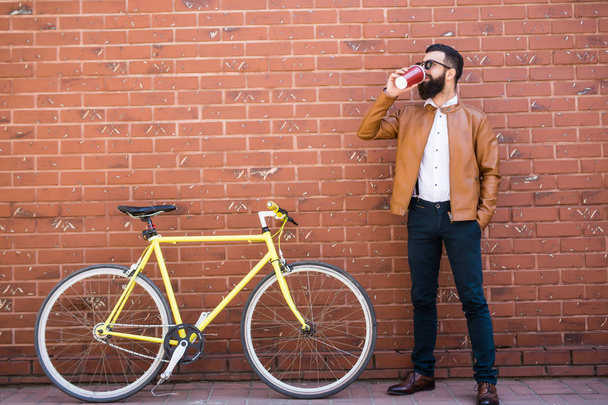 Молодий стильний чоловік з бородою сидить на цегляному велосипеді і п'є каву
 - Фото, зображення
