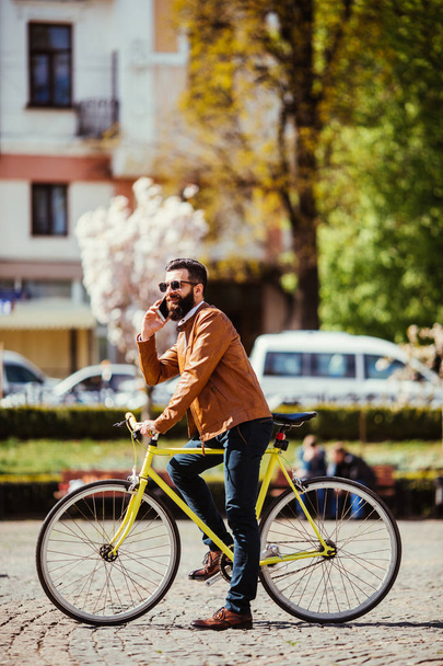 Cheerful young man with beard talking on the mobile phone and smiling while near his bicycle. - Photo, Image