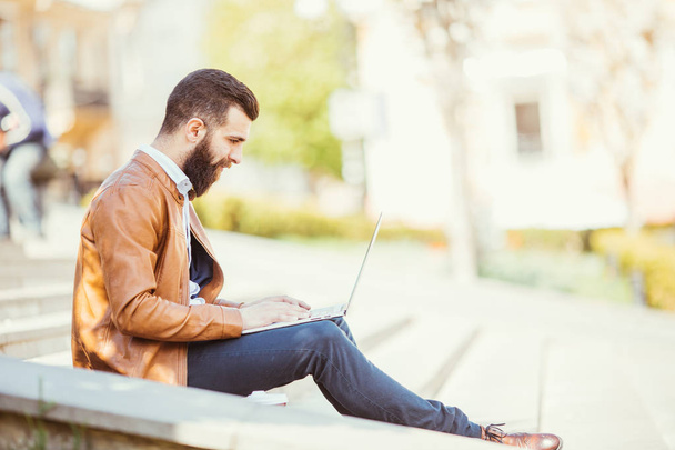 Young bearded handsome man sitting on the stairs using laptop in city - Photo, Image
