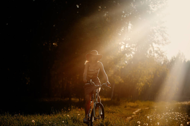 Забавная девушка водит велосипед на открытом воздухе. Солнечный летний образ жизни. Женщина в платье и шляпе в поле с одуванчиками. Женская прогулка в парке. Легкий фотоэффект для текста. Копировальное пространство для дизайна
. - Фото, изображение