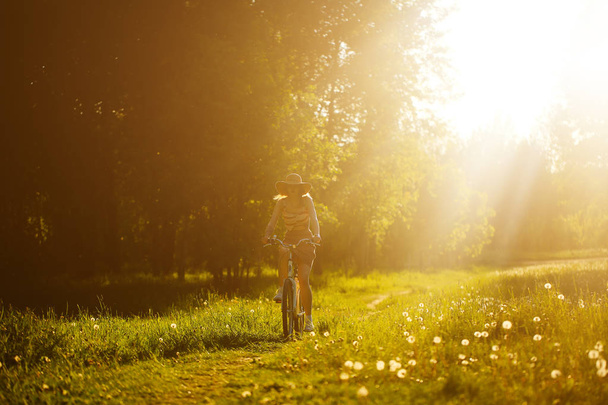 Забавная девушка водит велосипед на открытом воздухе. Солнечный летний образ жизни. Женщина в платье и шляпе в поле с одуванчиками. Женская прогулка в парке. Легкий фотоэффект для текста. Копировальное пространство для дизайна
. - Фото, изображение