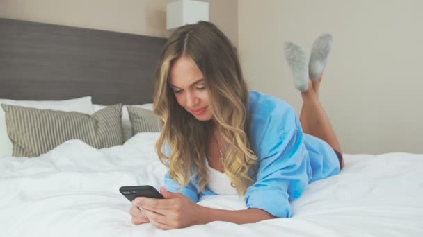 Beautiful woman lies on a bed on stomach, communicates on mobile phone - Video, Çekim