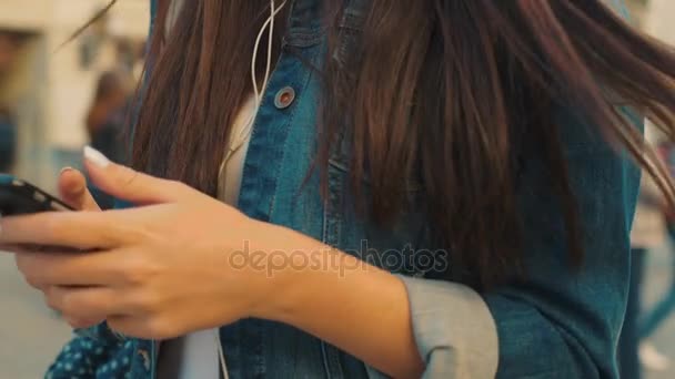 Beautiful young woman listening to music from her smart phone while walking on the street outside. Woman moving to the rythm. Happy woman using smart phone. Close up - Materiaali, video